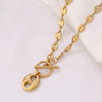 Collier Grains de Café en acier inoxydable doré, fermoir toggle, posé sur un tissu blanc et une pierre texturée, Maison Mercier.