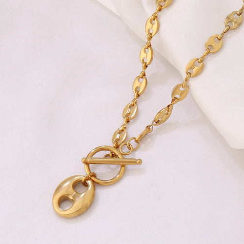 Collier Grains de Café en acier inoxydable doré, fermoir toggle, posé sur un tissu blanc et une pierre texturée, Maison Mercier.