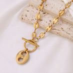 Collier Grains de Café en acier inoxydable doré, fermoir toggle, posé sur un tissu blanc et une pierre texturée, Maison Mercier.