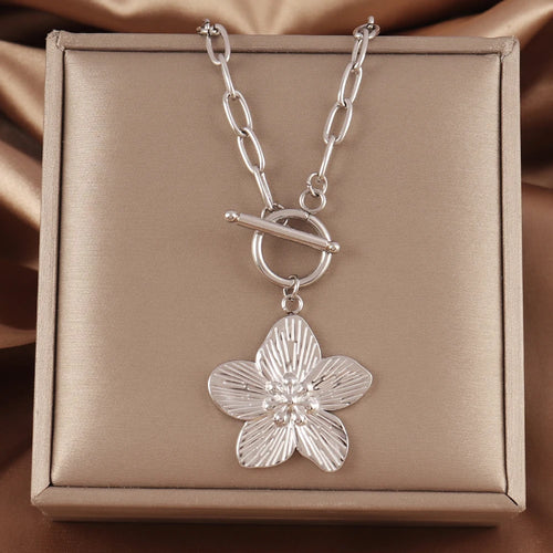 Collier chaîne ovale en acier inoxydable doré, avec pendentif fleur sculptée et fermoir T-Bar, présenté sur un écrin beige satiné, Maison Mercier.