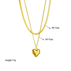 Quatre colliers dorés en acier inoxydable, présentant des pendentifs variés : un cercle noir, un cœur vert, un cœur avec une pampille noire, et un ensemble de chaînes avec étoiles et un pendentif carré bleu, présentés sur des supports beiges.