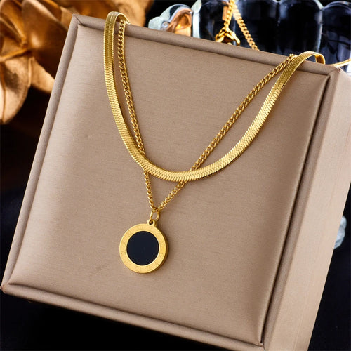Quatre colliers dorés en acier inoxydable, présentant des pendentifs variés : un cercle noir, un cœur vert, un cœur avec une pampille noire, et un ensemble de chaînes avec étoiles et un pendentif carré bleu, présentés sur des supports beiges.