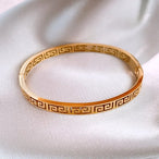 Détail d'un bracelet jonc rigide en acier doré, orné du motif Méandre Grec, présenté sur un drapé de soie blanche, Maison Mercier.