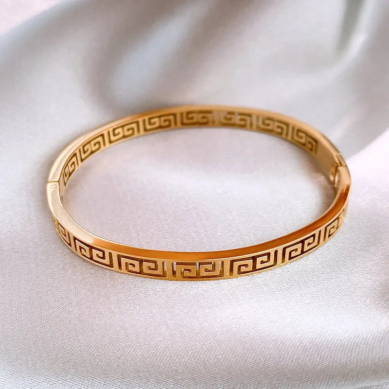 Détail d'un bracelet jonc rigide en acier doré, orné du motif Méandre Grec, présenté sur un drapé de soie blanche, Maison Mercier.