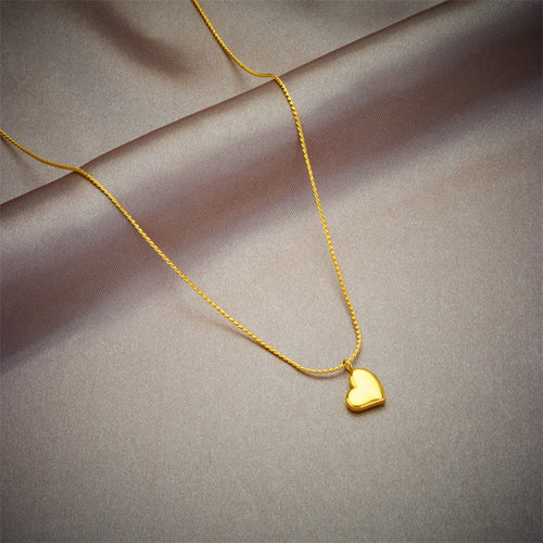 Détail d'un collier pendentif cœur 'Love' en acier inoxydable doré poli, posé sur un tissu élégant, Maison Mercier.