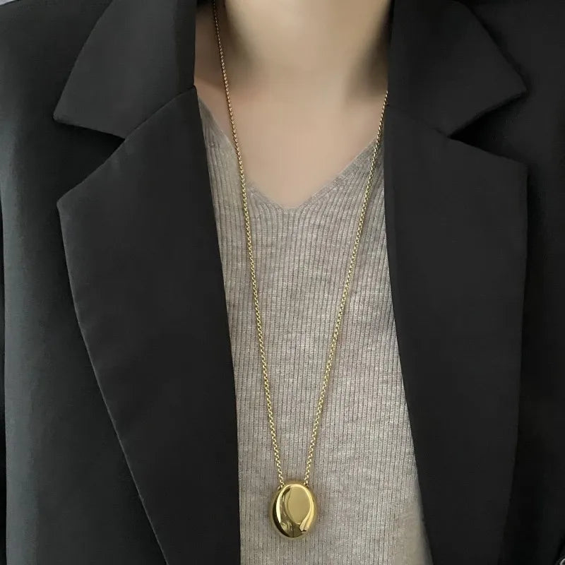 Deux colliers pendentifs ovales sculpturaux polis, l'un en finition argentée et l'autre en finition dorée, posés sur un tissu soyeux, Maison Mercier.