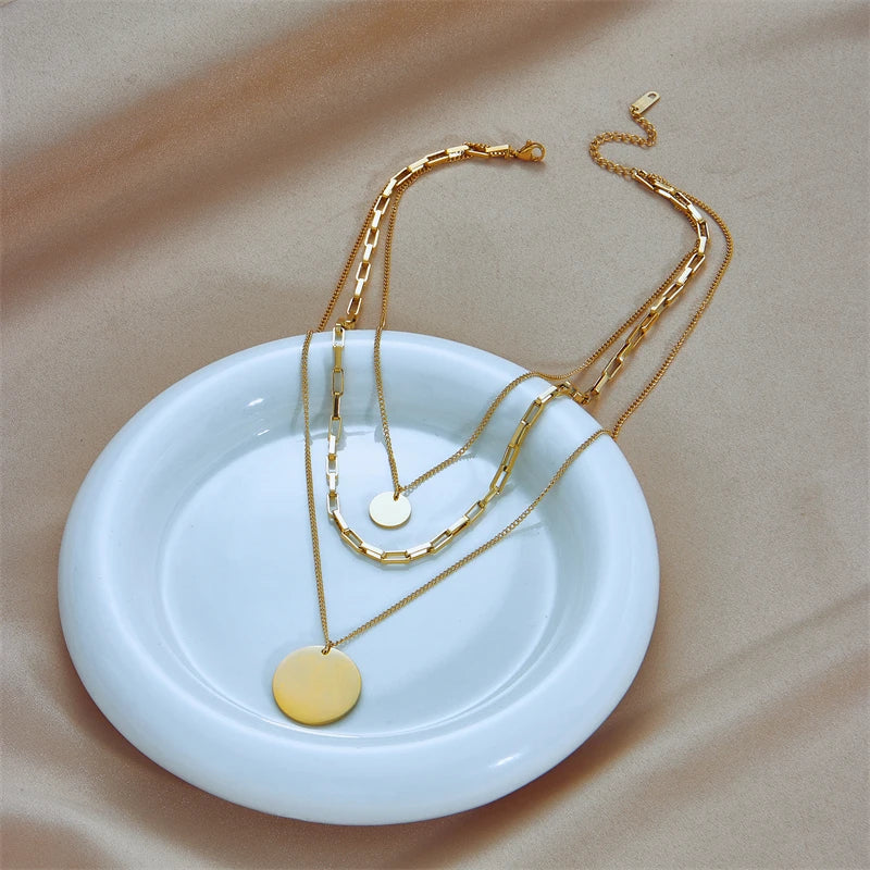 Collier multirangs en acier doré avec pendentifs disques, élégamment disposé sur une assiette en céramique blanche, Maison Mercier.