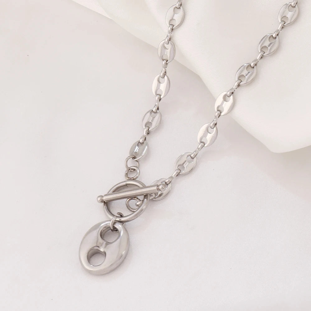 Collier Grains de Café en acier inoxydable doré, fermoir toggle, posé sur un tissu blanc et une pierre texturée, Maison Mercier.