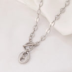 Collier Grains de Café en acier inoxydable doré, fermoir toggle, posé sur un tissu blanc et une pierre texturée, Maison Mercier.