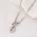Collier Grains de Café en acier inoxydable doré, fermoir toggle, posé sur un tissu blanc et une pierre texturée, Maison Mercier.