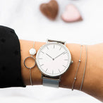 Montre féminine Maison Mercier, cadran blanc immaculé, bracelet maille milanaise en acier argenté, portée au poignet avec des bracelets argentés élégants, dont un avec un cercle et une pierre blanche.