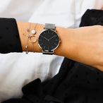 Montre féminine Maison Mercier, cadran noir minimaliste, bracelet maille milanaise en acier argenté, portée au poignet avec des bracelets fins argentés assortis.