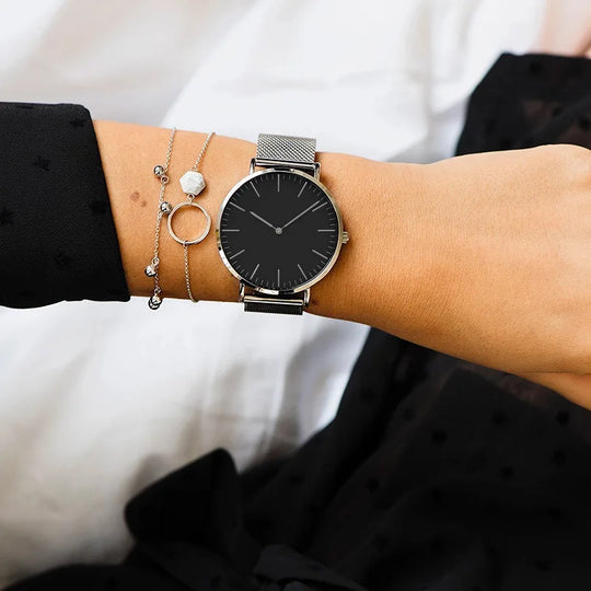 Montre féminine Maison Mercier, cadran noir minimaliste, bracelet maille milanaise en acier argenté, portée au poignet avec des bracelets fins argentés assortis.
