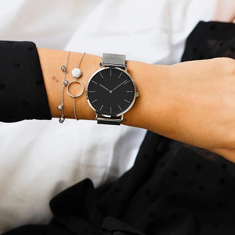 Montre féminine Maison Mercier, cadran noir minimaliste, bracelet maille milanaise en acier argenté, portée au poignet avec des bracelets fins argentés assortis.