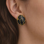 Détail d'une paire de boucles d'oreilles ovales en émail noir profond et finition or patiné, motif organique, posées sur un tissu soyeux rose pâle, Maison Mercier.