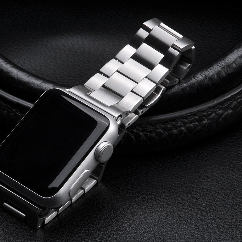 Détail d'un bracelet de montre à maillons classiques en acier inoxydable brossé argenté, fixé sur une montre connectée, posé sur un fond en cuir grainé, Maison Mercier.