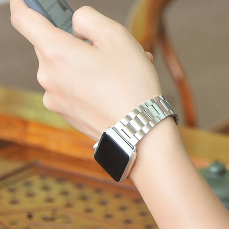 Vue d'un poignet portant un bracelet de montre à maillons classiques en acier inoxydable brossé argenté, la personne tenant un téléphone, dans un cadre lumineux et naturel, Maison Mercier.