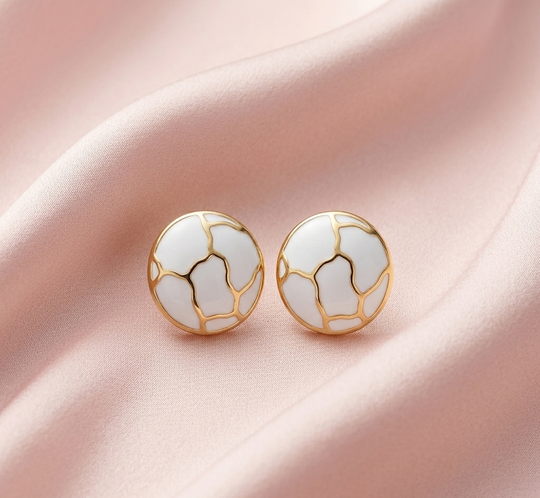 Détail d'une paire de boucles d'oreilles puces rondes en émail blanc et motifs dorés, posées sur un drapé de soie rose pâle, Maison Mercier.
