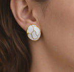 Détail d'une paire de boucles d'oreilles puces rondes en émail blanc et motifs dorés, posées sur un drapé de soie rose pâle, Maison Mercier.