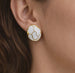 Détail d'une paire de boucles d'oreilles puces rondes en émail blanc et motifs dorés, posées sur un drapé de soie rose pâle, Maison Mercier.