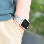 Gros plan d'un poignet portant un bracelet de montre à maillons classiques en acier inoxydable brossé argenté, la personne vêtue d'un polo bleu et d'un pantalon beige, en extérieur, Maison Mercier.