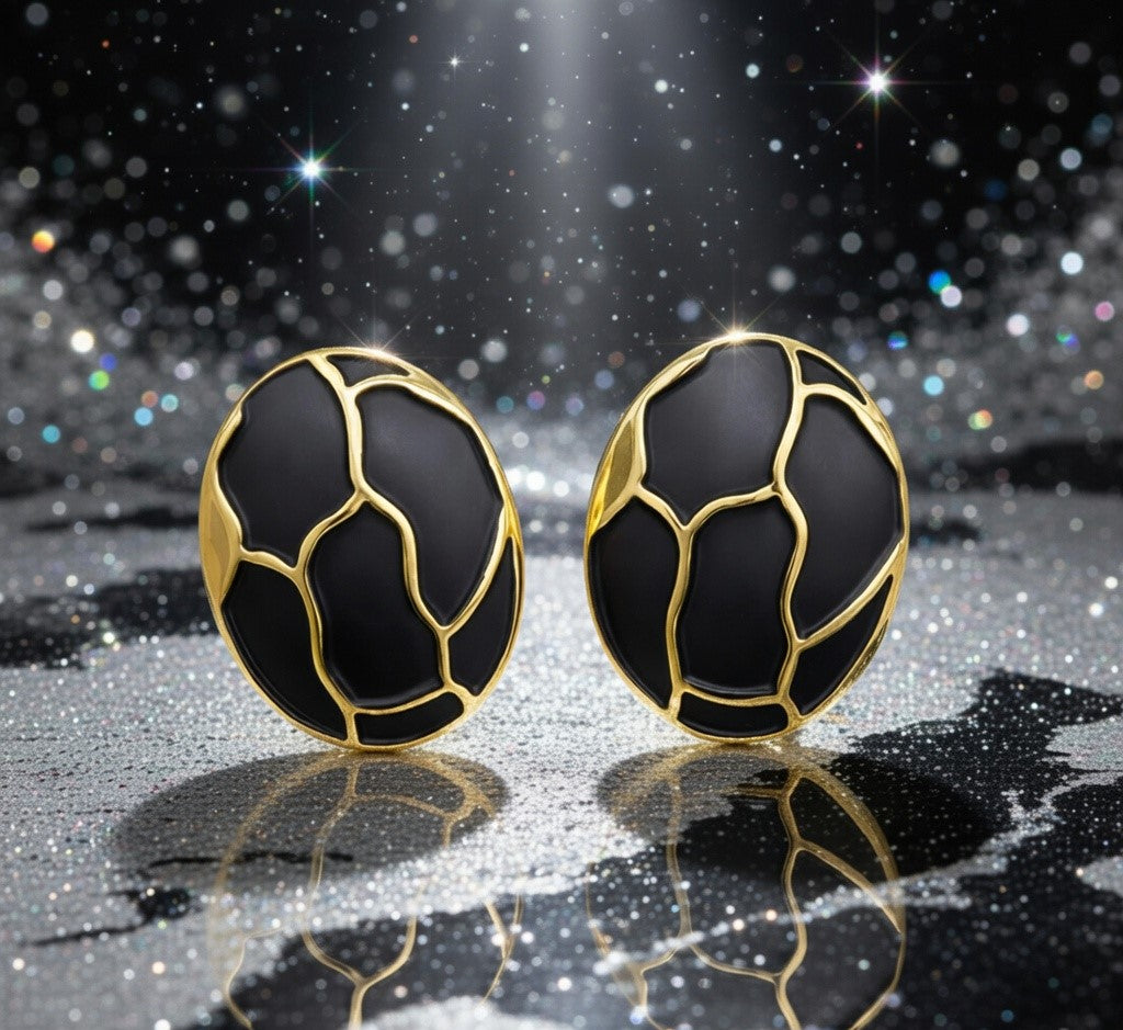Détail d'une paire de boucles d'oreilles ovales en émail noir profond et finition or patiné, motif organique, posées sur un tissu soyeux rose pâle, Maison Mercier.