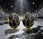 Détail d'une paire de boucles d'oreilles ovales en émail noir profond et finition or patiné, motif organique, posées sur un tissu soyeux rose pâle, Maison Mercier.