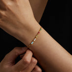 Détail d'un bracelet chaîne fine plaqué or, orné de six pierres de naissance rectangulaires multicolores, posé sur un fond texturé clair.