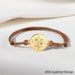 Bracelet cordon ajustable avec médaillon plaqué or finement gravé de motifs floraux et célestes, présenté sur un tissu blanc immaculé, soulignant la délicatesse de la gravure.