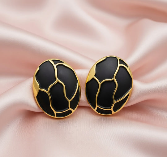 Détail d'une paire de boucles d'oreilles ovales en émail noir profond et finition or patiné, motif organique, posées sur un tissu soyeux rose pâle, Maison Mercier.