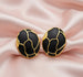 Détail d'une paire de boucles d'oreilles ovales en émail noir profond et finition or patiné, motif organique, posées sur un tissu soyeux rose pâle, Maison Mercier.