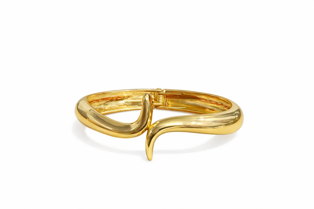 Détail d'un bracelet jonc à la finition dorée éclatante, présentant une esthétique de vague fluide et un fermoir articulé, Maison Mercier.