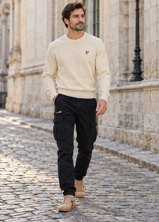 Un homme adulte portant Pull Lyle & Scott beige homme, Pantalon Replay noir homme, Baskets Diadora Game Low Suede homme. Le pavé sous les pas. Mise en scène IA illustrative.