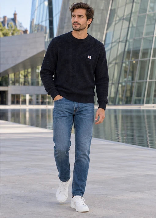 Un homme adulte portant Pull Calvin Klein Jeans noir homme, Jean Tommy Hilfiger Denton Slim Straight homme, Baskets Calvin Klein Jeans blanc homme. Devant le musée. Mise en scène IA illustrative.